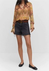 Blusa a maniche lunghe a fantasia in arancione, giallo e verde, abbinata a pantaloni corti in denim neri con orli sfilacciati e una cintura marrone. Ballerine nere.