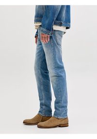 Πλάγια όψη ατόμου που φοράει ανοιχτό μπλε τζιν, ταμπά σουέτ μποτάκια, τζιν τζάκετ και κρεμ πουκάμισο σε απλό λευκό φόντο.