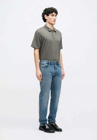 Πόλο μπλούζα με κοντά μανίκια σε ελιά πράσινο με διακριτικό μοτίβο, συνδυασμένη με ανοιχτόχρωμα τζιν μπλε και μαύρα δερμάτινα παπούτσια. Το μοντέλο στέκεται πλαγίως.