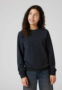 Giovane donna con capelli ricci, che indossa una maglietta scura a maniche lunghe e jeans blu, che guarda in alto e sorride leggermente davanti a uno sfondo neutro.