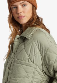 Quiltet grøn jakke med spidse krave, stor forlommek og brun strikket beanie. Stof ser letvægtigt og struktureret ud.