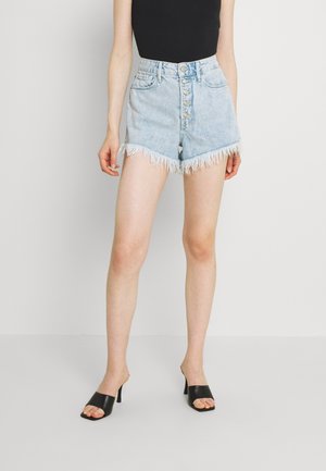 Vrouw draagt lichtblauwe high-waist denim shorts met een rafelige zoom en knopen aan de voorkant, een zwart mouwloos topje en zwarte sandalen met hak.