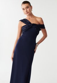 Nocna suknia w kolorze granatowym z twisted, beaded dekoltem na jedno ramię, dopasowanym kroju i bocznym rozcięciem dla swobody ruchów.