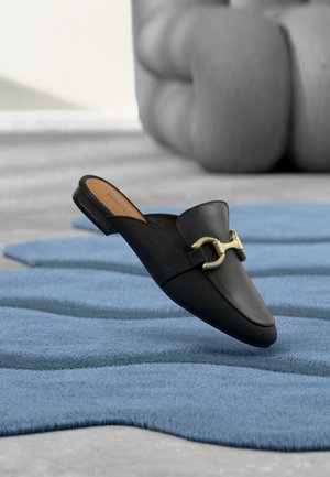 Mocassin en cuir noir sans lacets avec détail mors doré reposant sur un tapis bleu texturé, art sculptural gris en arrière-plan flou.