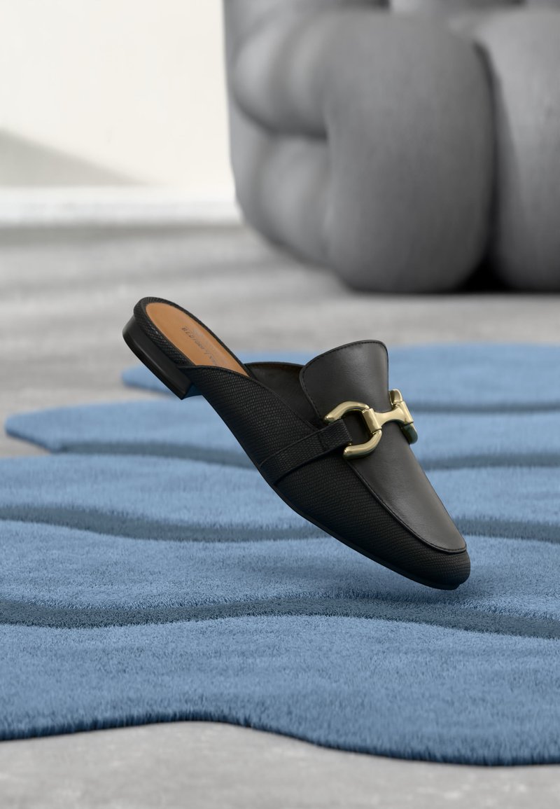 Mocassin en cuir noir sans lacets avec détail mors doré reposant sur un tapis bleu texturé, art sculptural gris en arrière-plan flou.