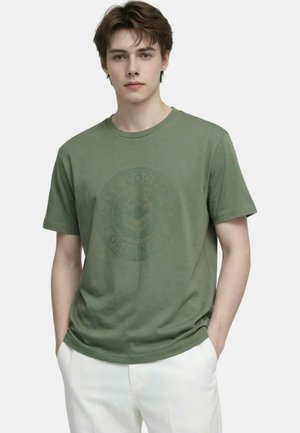 Jeune homme portant un t-shirt graphique vert olive et un pantalon blanc, debout les mains dans les poches devant un fond uni.