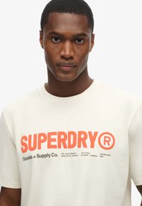 T-shirt en coton crème avec un grand logo rouge "SUPERDRY", accompagné de texte plus petit et de détails de marque en dessous. Design classique à col rond.