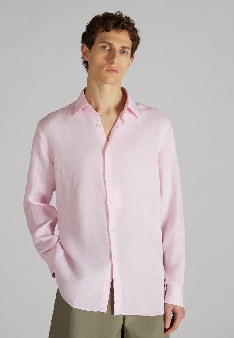 Joven con cabello rizado que lleva una camisa de manga larga rosa claro desabotonada y pantalones verde oliva, de pie contra un fondo gris liso.