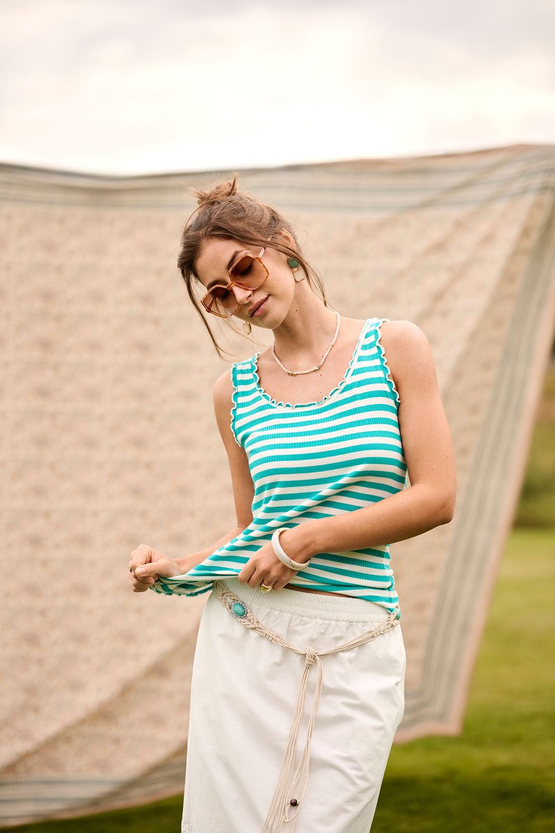 Femme portant un débardeur turquoise à rayures et une jupe blanche ajuste sa chemise en extérieur avec un fond flou en tissu à motifs.