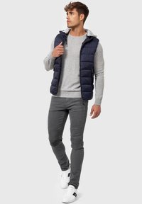 Marineblå puffer vest over en lysegrå sweater, parret med grå slim-fit jeans med ribbede detaljer og hvide sneakers med sorte detaljer.