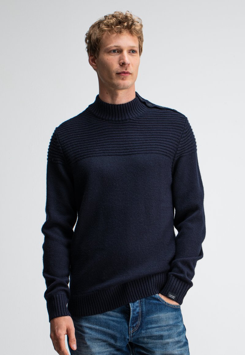 Man met krullend blond haar, gekleed in een donkerblauw ribgebreid trui en blauwe jeans, staand met één hand in de zak tegen een effen achtergrond.