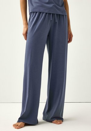 Persoon draagt losse, blauw wijde broek met aantrekkoord in de taille, staat blootsvoets op een lichte vloer tegen een effen achtergrond.