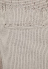 Tissu beige avec de fines rayures blanches verticales, montrant une poche passepoilée cousue et une ceinture élastique au-dessus.
