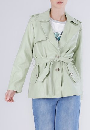 Donna con un trench color verde chiaro con cintura e grandi revers, tasche con bottoni, indossato sopra una maglietta bianca a fiori e jeans blu, in piedi con un braccio abbassato.