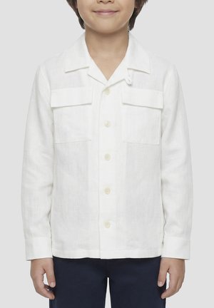 Enfant portant une chemise blanche à manches longues avec deux poches poitrine, debout devant un fond clair uni.