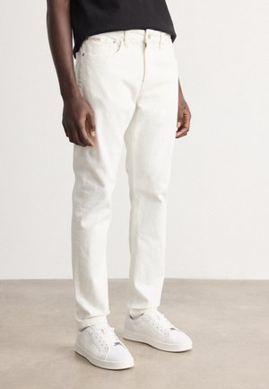 Homme portant un jean blanc droit et des baskets blanches, debout sur un sol clair contre un mur uni.