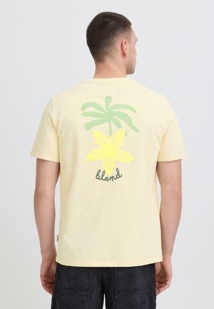 Mann med kort hår iført en lys gul t-skjorte med et design av en grønn palme og en gul blomst, med ordet «blend» på baksiden.