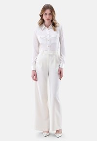 Blusa di seta bianca con due tasche frontali, chiusa con bottoni; abbinata a pantaloni bianchi a vita alta e a gamba larga. Gli accessori includono tacchi a punta bianchi.