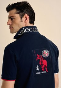 Polo de color navy con un cuello alzado, texto "valecuatro", logo de jinete rojo bordado y terminaciones rojas en las mangas.