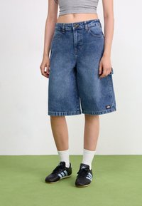 Bermudas de mezclilla en azul con un corte holgado, cinco bolsillos y dobladillo enrollado. Combinadas con zapatillas deportivas negras y calcetines blancos.