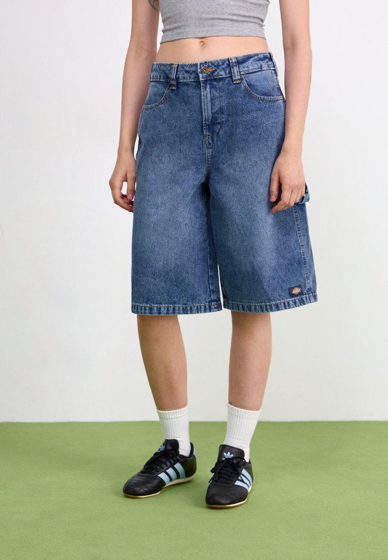 Bermudas de mezclilla en azul con un corte holgado, cinco bolsillos y dobladillo enrollado. Combinadas con zapatillas deportivas negras y calcetines blancos.