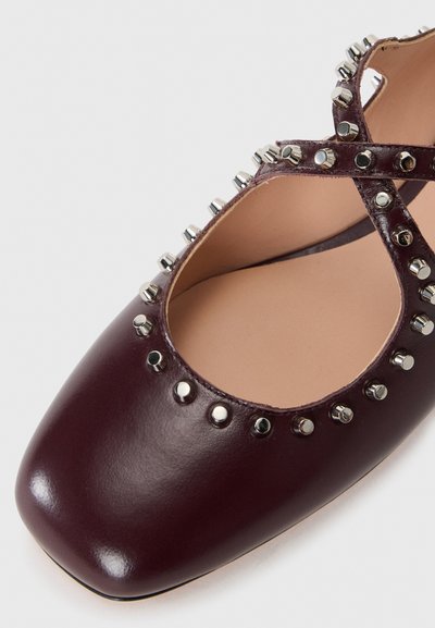 Bailarina de cuero burdeos con punta cuadrada, adornada con tachuelas metálicas alrededor de la abertura y un diseño de tiras cruzadas. Textura suave.