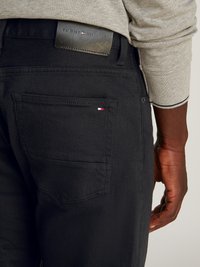 Juodų deniminių kelnių su odiniu Tommy Hilfiger juosmens žymeniu. Yra užpakalinis kišenė su mažomis raudonos, baltos ir mėlynos spalvų logotipo akcentais.