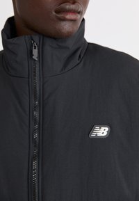 Svart lättviktsjacka med hög krage och dragkedjestängning. Har en texturerad tyg och en liten vit New Balance-logotyp på bröstet.