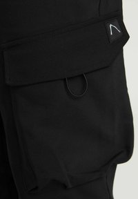 Un pantalon cargo noir présente une grande poche latérale avec un rabat, des détails en boucle élastique et un tissu lisse et texturé, soulignant la praticité.
