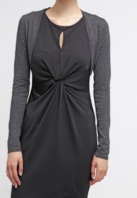 Schwarzes Kleid mit gedrehtem Frontdetail und Schlüsselloch-Ausschnitt, kombiniert mit einer taillierten grauen Strickjacke mit langen Ärmeln. Glattes, elegantes Material.