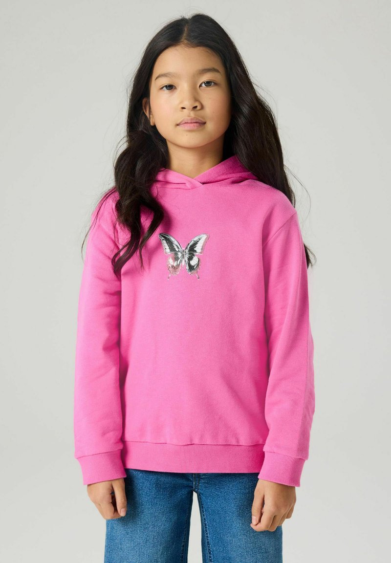 Jeune fille aux longs cheveux foncés portant un sweat à capuche rose vif avec un motif de papillon noir et blanc et un jean bleu, sur un fond clair.