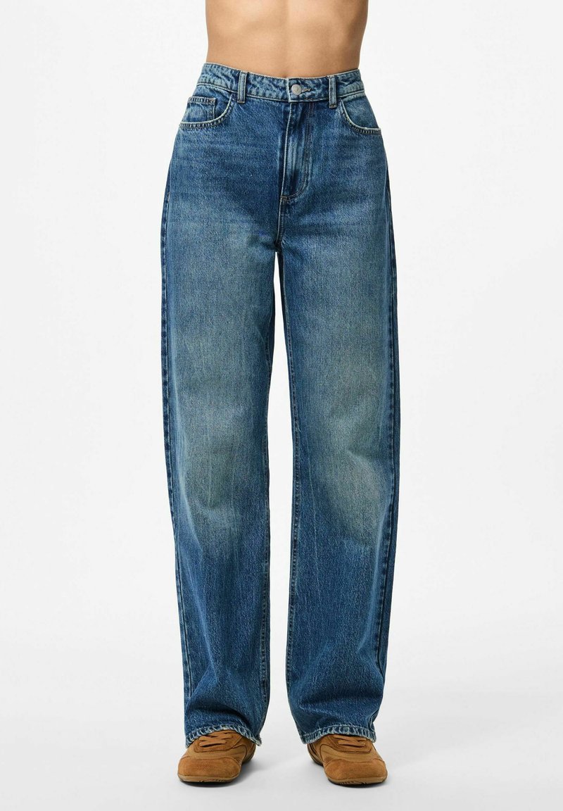 Široke plave traperice s izblijedjelim završetkom, visokim strukom, prednjim džepovima i šivanjem na rubu. Uparene s smeškim nubuk cipelama.