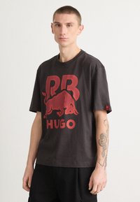 Black t-shirt featuring a large red bull graphic and "HUGO" text. It has a relaxed fit, short sleeves, and a tag on the sleeve.