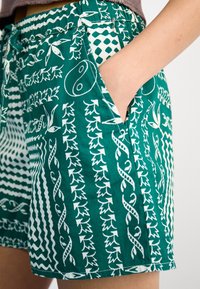 Pantaloni scurți verzi cu modele albe, prezentând frunze și desene abstracte. Talie elastică și buzunare pe ambele părți. Material din bumbac.