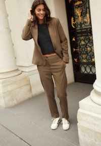 Costume marron ajusté avec un blazer cintré et un pantalon droit, associé à un pull court noir et des baskets blanches.
