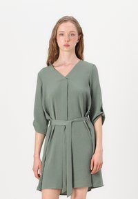 Vestido verde, até ao joelho, com decote em V, mangas três quartos e um cinto atado na cintura. Tecido texturizado com um acabamento suave.