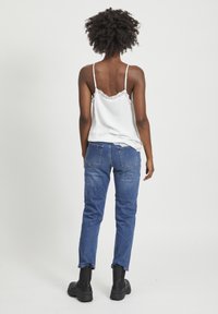 Weiße Camisole mit Spitzenrand und dünnen Trägern, kombiniert mit blauen Jeans und schwarzen Knöchelstiefeln, von hinten betrachtet.