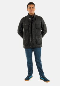 Zwarte nylon jas met vier voorkruiken en drukknoopsluitingen, gecombineerd met blauwe denim jeans en sneakers met blauwe strepen en rubberen zolen.