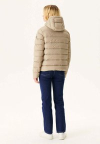 Chaqueta acolchada beige con diseño de rombos horizontales, capucha adjunta y parche con el logotipo. Combinada con jeans azul oscuro y zapatillas blancas.