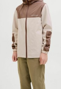 Giacca con zip beige chiaro e marrone con cappuccio, caratterizzata da sezioni a blocchi di colore e polsini elastici. Modello con pantaloni verde oliva.