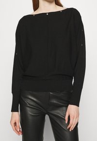 Sort oversized sweater med bred halsudskæring, ribbede manchetter og kant. Har sølvfarvede trykknapper på skuldrene. Fremstillet af et blødt, struktureret stof.