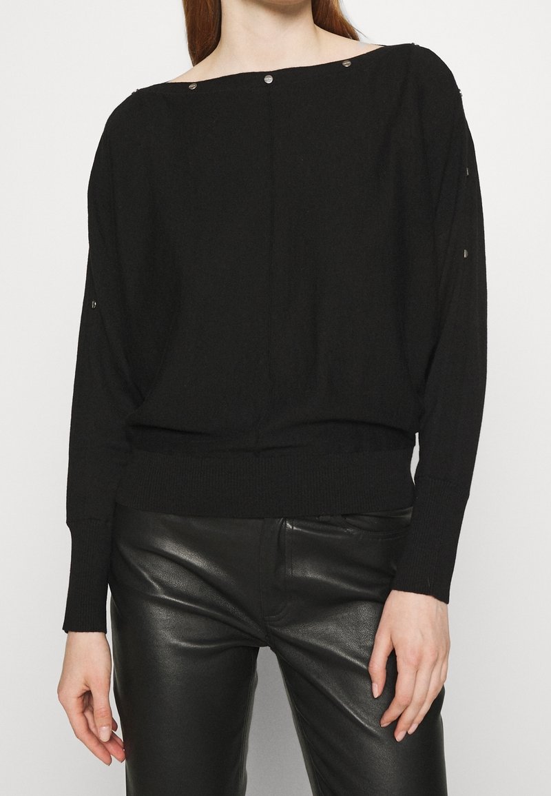 Sort oversized sweater med bred halsudskæring, ribbede manchetter og kant. Har sølvfarvede trykknapper på skuldrene. Fremstillet af et blødt, struktureret stof.