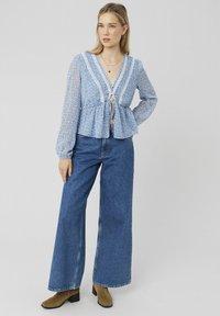 Blouse florale bleu clair avec une taille peplum, bordure en dentelle blanche, devant noué, manches longues en tissu transparent, accompagnée de jeans bleus à jambes larges et de bottines marron.
