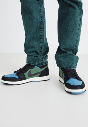 Zapatillas con paneles negros, verdes y azules, usadas con vaqueros texturizados color verde azulado sobre un suelo blanco.