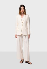 Completo su misura color crema con giacca doppiopetto e pantaloni a gamba larga, abbinato a sandali neri e un top a scollo a V sottostante.