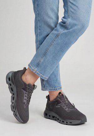 Beine in blauen Jeans, die schwarze Sportschuhe mit dicken Sohlen und Belüftungslöchern tragen, stehend vor einem schlichten hellen Hintergrund.