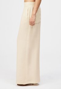 Beżowe szerokie spodnie z wysokim stanem, elastycznym pasem i bocznymi kieszeniami, noszone z brązowymi sandałami na obcasie z odkrytymi palcami.