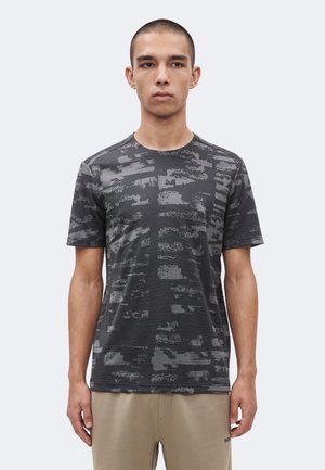 Noor mees, kes kannab musta ja halli mustriga lühikest T-särki ja beežid pükse, seisab ühevärvilise heleda tausta ees.