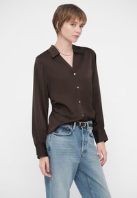 Lühikarvaline naine kannab tumeda pruuniga nööbitud pluusi, mis on sisse pandud helesinistesse kõrge vöökohaga teksadesse ja musta vööd, valge tausta ees.