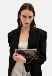Pochette in metallo con finitura lucida, forma rettangolare, tracolla staccabile, tenuta in mano su uno sfondo di blazer nero.